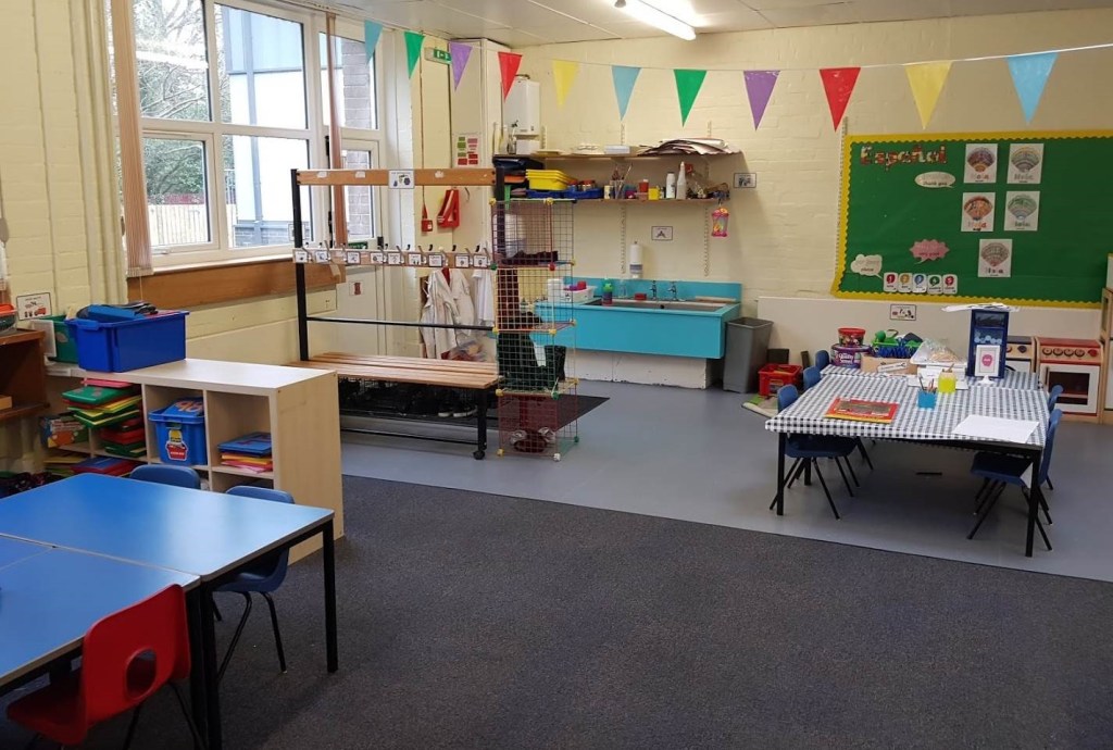 the primary one classroom before, there are two banks of rectangular tables and plastic blue chairs, a cloakroom bench takes up space in the classroom.