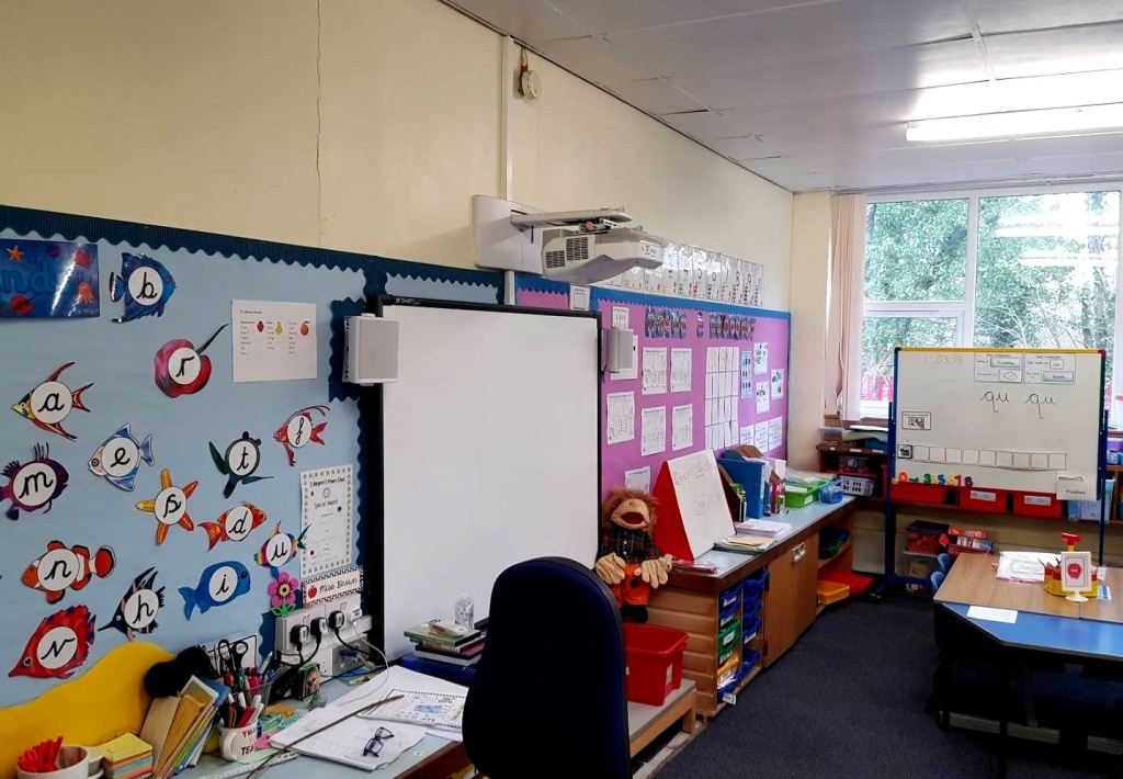 the primary one classroom before, the teaching wall display is blue and purple, the storage is wooden with plastic blue and green trays, there is a table with chairs beside a whiteboard