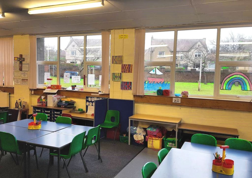 the classroom has yellow walls, blue tables and green plastic chairs, the room appears dark although there are a lot of windows.