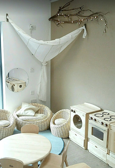 A home corner with wicker seating, wooden tables and chairs and wooden play kitchen equipment at Longstone Primary School Nursery
