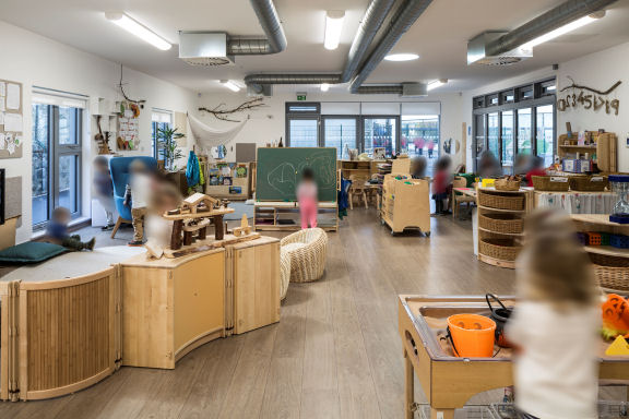 a child plays in the sand box, another is drawing on a chalk board, a group of children play in a comfy corner. One child play with the dolls house and a group is doing an activity with a member of staff