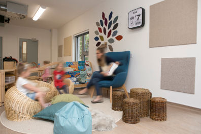 a library corner where a member of staff reads to two children from an armchair, the children are sitting in front on wicker chairs