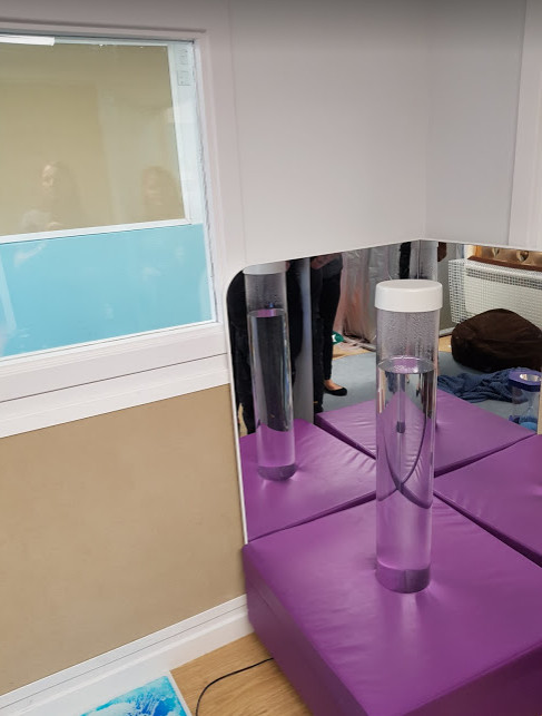 a sensory bubble tube is inset on a square purple cushioned base with a mirror curving around two sides of the cushion