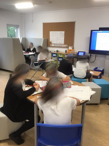 primary seven children sitting at different areas of the classroom. three sit at a round table, two sit on upholstered cubes, one child sits on his own at a chair with a flip down desk, a group sit in a booth