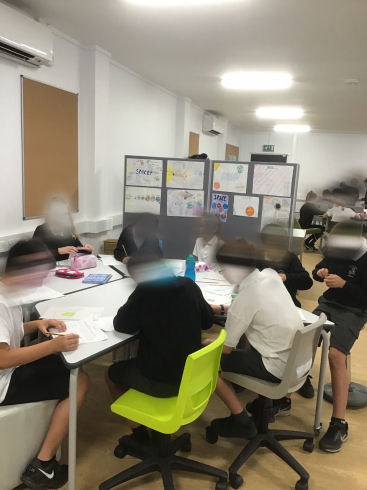 primary seven children sitting together at flexible tables. some on wheeled chairs, one on a high stool another on a cube