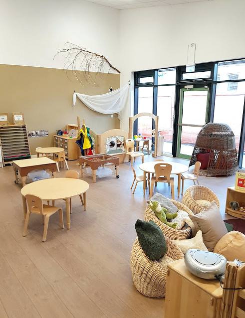 the classroom with large windows from the floor, wicker basket chairs and cushions. three round tables with chairs, a wicker nook and sand and water play units.