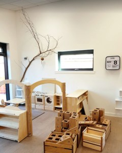 a home corner with play kitchen is accessed through a little archway, outside of the partitioned area are lots of blocks for construction. A branch hangs on the wall