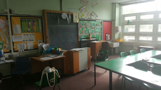 the teaching wall has a blackboard and teachers desk, the room is bright with natural daylight but the furniture and carpet darkens the room