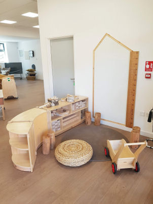 a giant ruler is on the wall beside a house shaped white board, a wicker seat pad is on a rug on the floor beside the block play storage that is low down for children to access