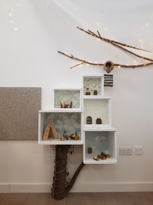 nature inspired decoration, this shelving unit has a tree trunk from the floor to the base of the unit, a branch with fairy lights is hanging on the wall above