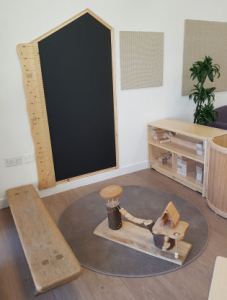 A giant wooden ruler is on the wall beside a house shaped blackboard for children to write on. a bench is beside a dolls house which is sitting on a circular rug