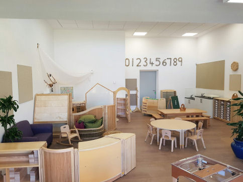 in the classroom there are water and sand play units, areas are partitioned off using low down shelving units, there is a circular table and chairs and numbers on the wall