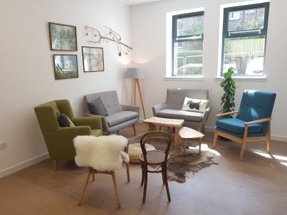 an adult area that looks like a living room with a mixture of different armchairs and wooden chairs in a circle, coffee table and a rug in the centre and a lamp in the corner