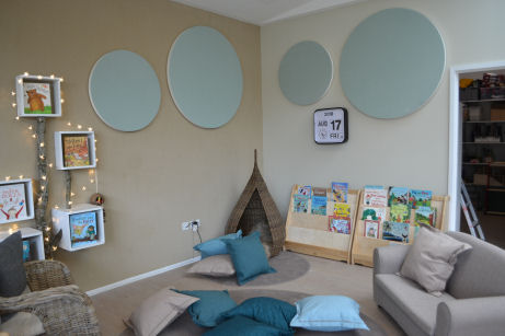 the reading corner looks like a living room with a sofa, lots of cushions, rugs and a wicker teacher's chair