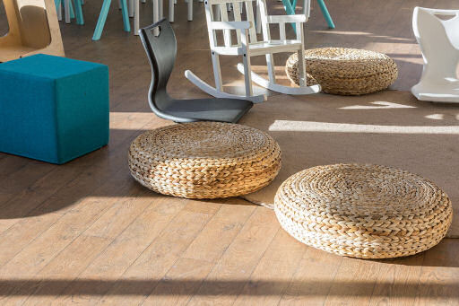 On the floor of the classroom surrounding a circular rug are three wicker seat pads, an upholstered blue cube seat, a plastic floor seat which has a high back support and no legs and rocks and a white rocking chair.
