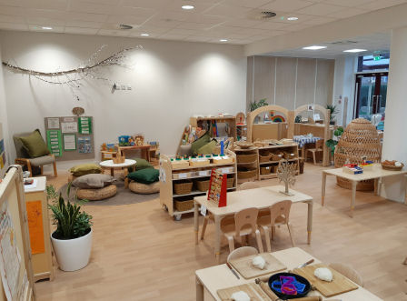 the classroom with lots of natural decoration, wooden tables, chairs and storage units, plants are placed around the room, baskets, floor pads and a hideaway nook are made from wicker, there is a branch with fairy lights hanging on the wall