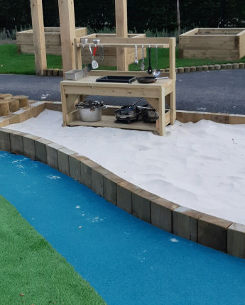 in the outdoor play area there is artificial grass, a wavy blue wetpour surface has been designed around the sandpit to look like water. a mud kitchen is inside the sandpit