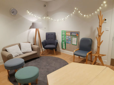 an adult seating area is decorated like a lounge with a hexagonal wooden table, upholstered circular stools, a sofa, lamp and armchairs