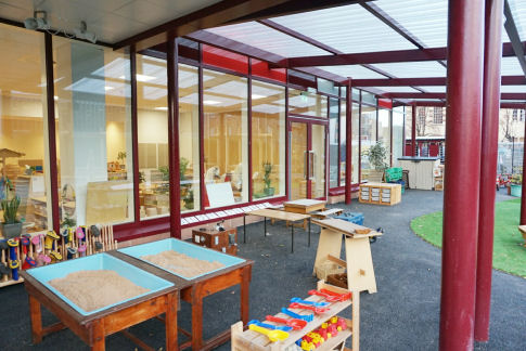 the outdoor space underneath a canopy where there is sand play units and a workbench, there is a welly rack. The space has been designed like an outdoor classroom
