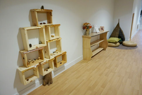 wooden box shelving on the wall is shaped like a tree with a trunk at the bottom going down to the floor. a wooden bookshelf sits next to a cozy reading area with a wicker teepee and seat pads resting on a circular rug