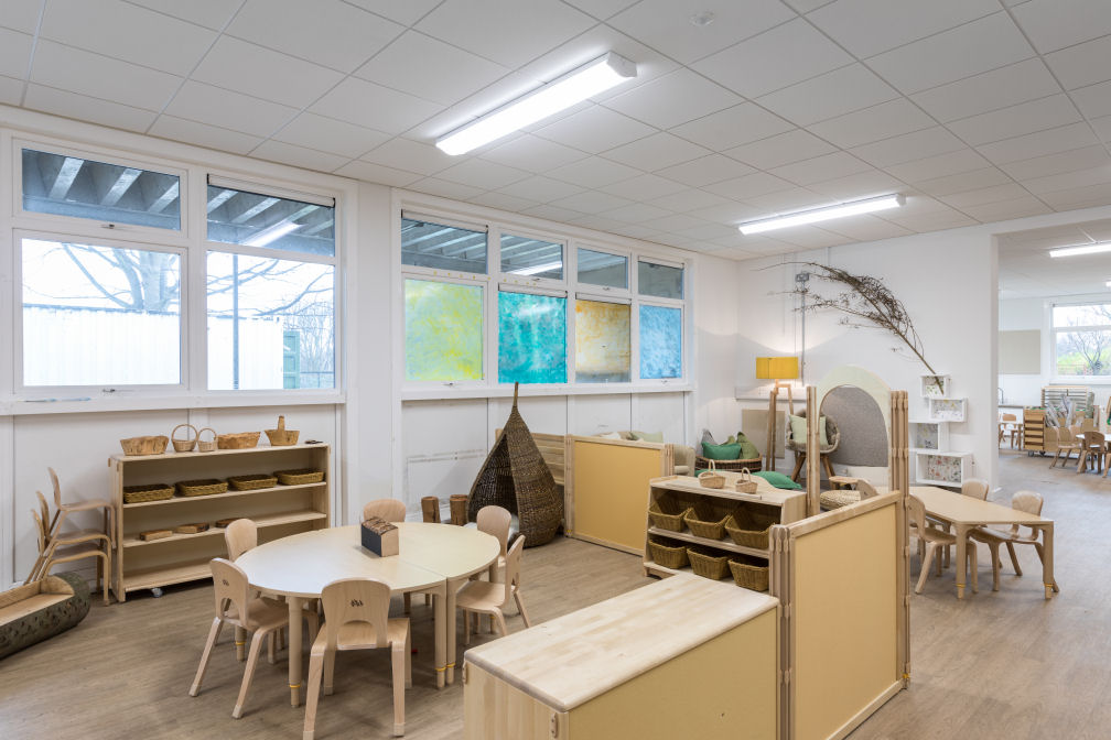 the nursery room is decorated in natural wooden colours for the flooring, tables, chairs and storage units. There is a wicker nook to sit in beside a round table with six small chairs. On the floor is a small bench carved from a log. There is a tree branch on the wall.