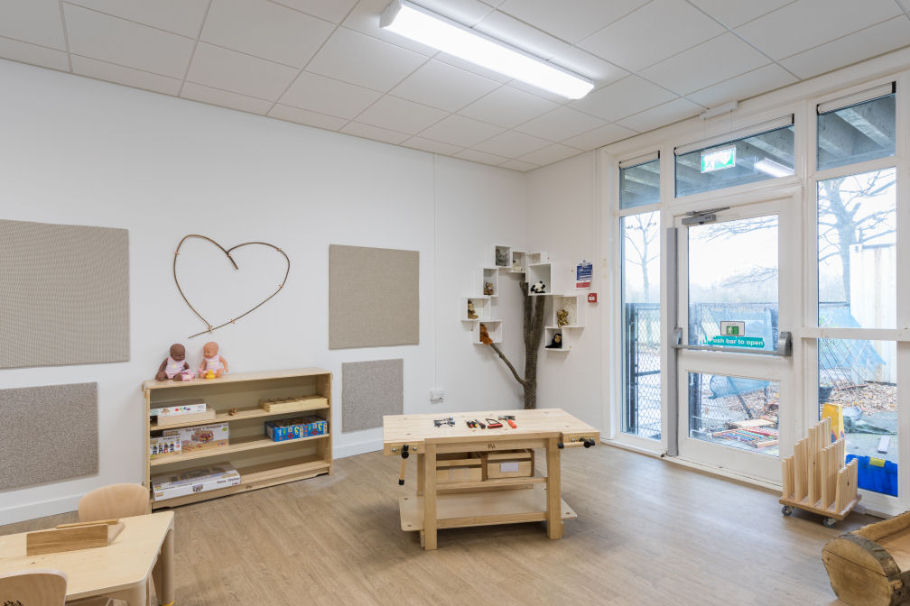 the walls are white and the flooring and furniture are wooden. There is a decorative heart on the wall and a real tree trunk with branches in the corner that has white cube shelves surrounding it to look like the foliage. There are fabric pin boards on the wall. In the centre is a child's workbench. There is a wooden welly stand next to the door, the glazing is floor to ceiling so you can see out to the playground.