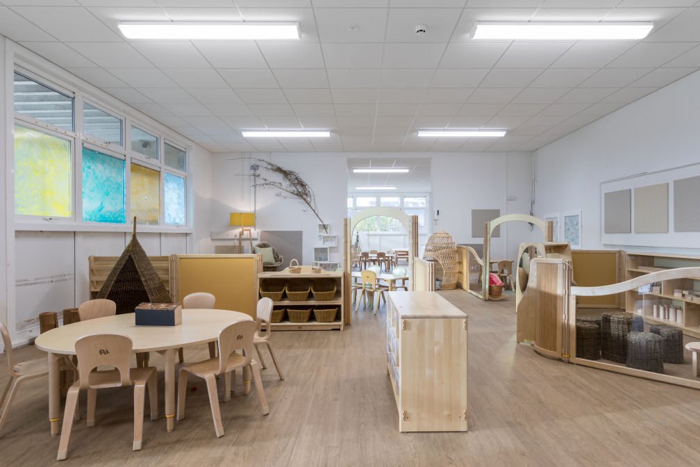 the nursery room is decorated in natural wooden colours for the flooring, tables, chairs and storage units. There is a wicker nook to sit in behind a round table with six small chairs. There is a tree branch on the wall. The room is separated into zones using dividers and archways, some of the dividers are solid and some are see through. There are wicker stools in a group on the floor.