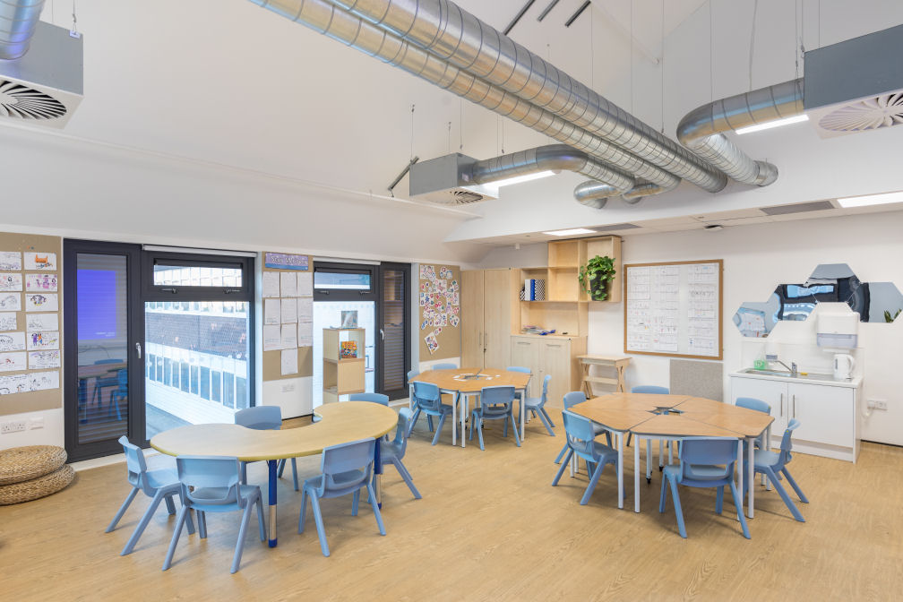 This room is decorated in natural wood colours for the floor, tables and storage units. There are stone coloured plastic chairs at the tables. Two of the tables can split up into six individual triangular tables and one is a U shape. there are hexagon mirrors on the wall, a plant on a shelf and beige pin boarding displaying children's work