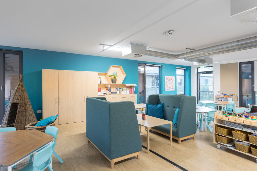 A classroom with floor to ceiling windows, natural wood colours for the floor, tables and storage units with a blue wall and coordinating blue chairs and cushions. In the centre are two soft upholstered blue booths facing each other over a table. There is a wicker nook to sit in by the wall.