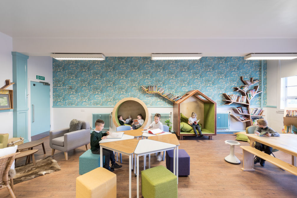 One wall has blue wallpaper printed with flowers the other wall is painted white with blue accents. Against one wall are two large wooden nooks to sit in. Two children sit facing each other to read in the circular nook. One child sits on a large cushion in the larger house shaped nook and reads a book. On one side of the room is a living room set up with an armchair, rug and coffee tables. In the middle of the room, two boys sit at a table that can split into six individual triangular tables on colourful upholstered cube stools and a plastic chair. Another boy sits in a comfy chair at a large wooden dining table.