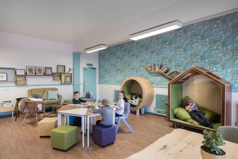 One wall has blue wallpaper printed with flowers the other wall is painted white with blue accents. Against one wall are two large wooden nooks to sit in. Two children relax to read in the circular nook, resting their backs on cushions. One child lies down on a large cushion in the larger house shaped nook and reads a book. At the far side of the room is a living room set up with an armchair, sofa and wicker chairs in a circle with little coffee tables. In the middle of the room, three boys sit at a table that can split into six individual triangular tables on colourful upholstered cube stools and a plastic chair.