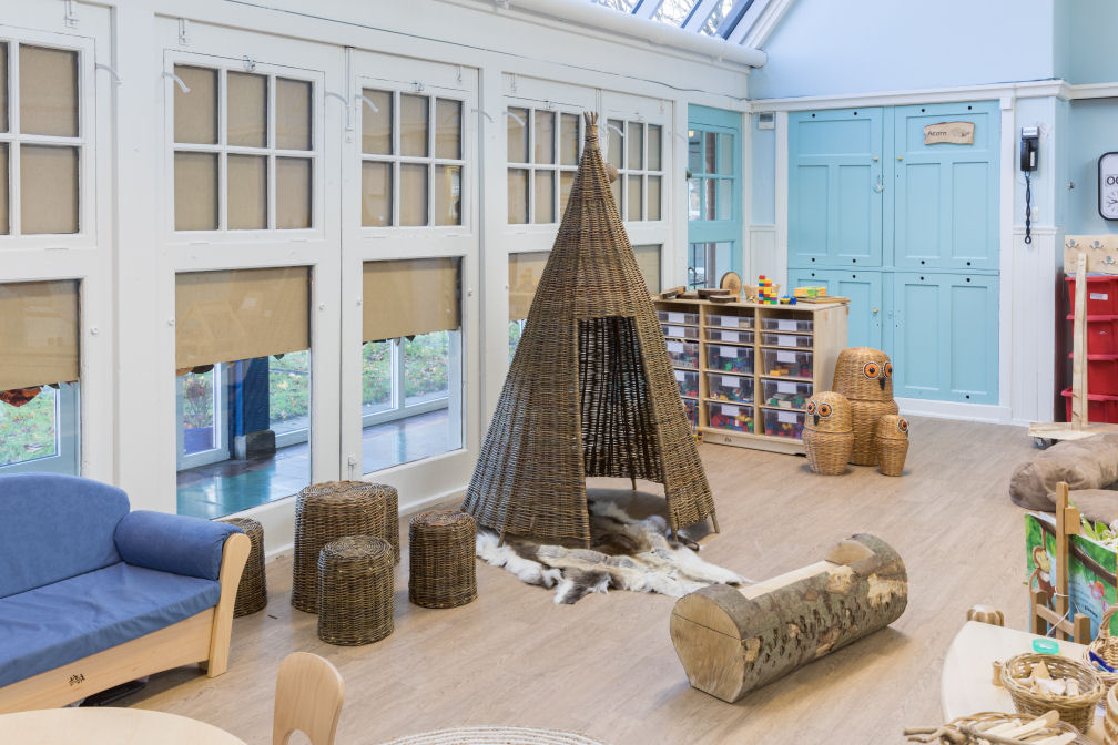 In the centre is a tall tipi style wicker nook, surrounded by a group of different sized wicker stools and a little bench carved from a log. Beside this is a blue sofa and at the rear of the room is a storage unit fulled with colourful blocks and three wicker owl shaped baskets.