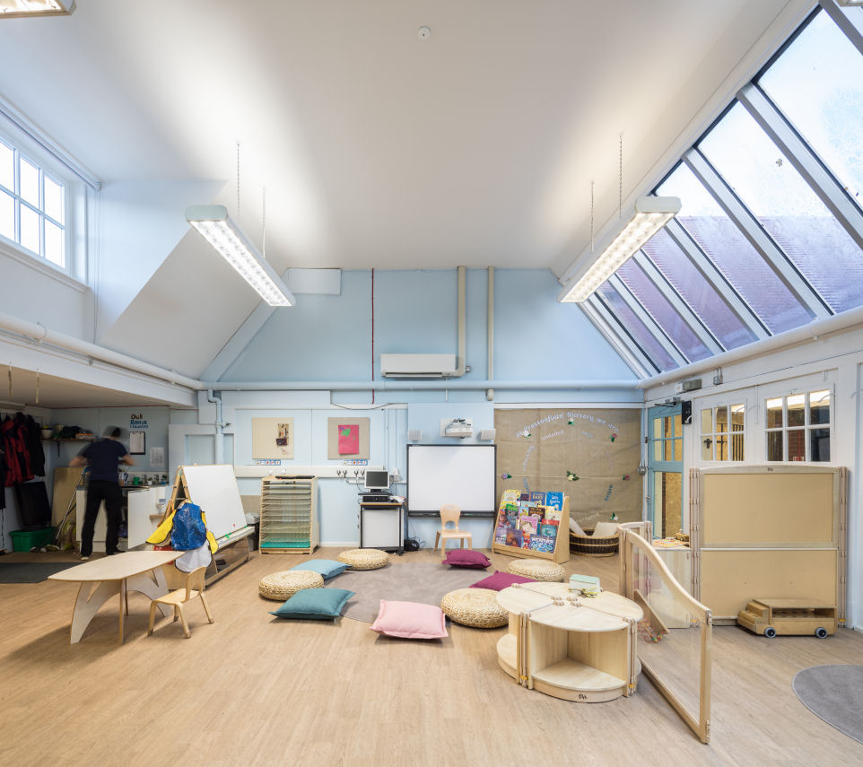 The nursery room is bright with natural light. there are natural wooden furnishings and the flooring is a wooden colour. In the centre on a pale blue wall is a Smart board and laptop on a trolley. There is a hessian fabric display board. In front of the Smart board a circular rug is surrounded by pink and blue cushions and wicker seat pads.