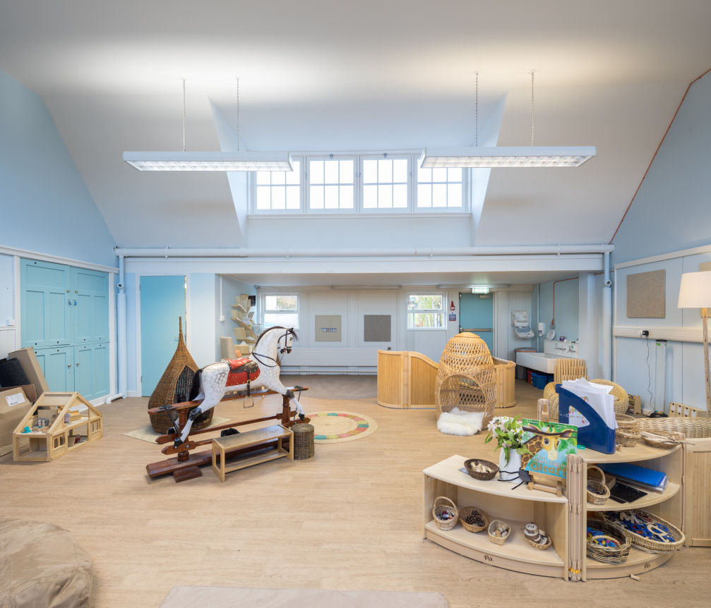 In this room are natural wooden furnishings and the flooring is a wooden colour. In the centre of the room is a traditional style rocking horse, a circular rug and two different wicker nooks. At the front are open shelving units with wicker baskets that show the contents easily