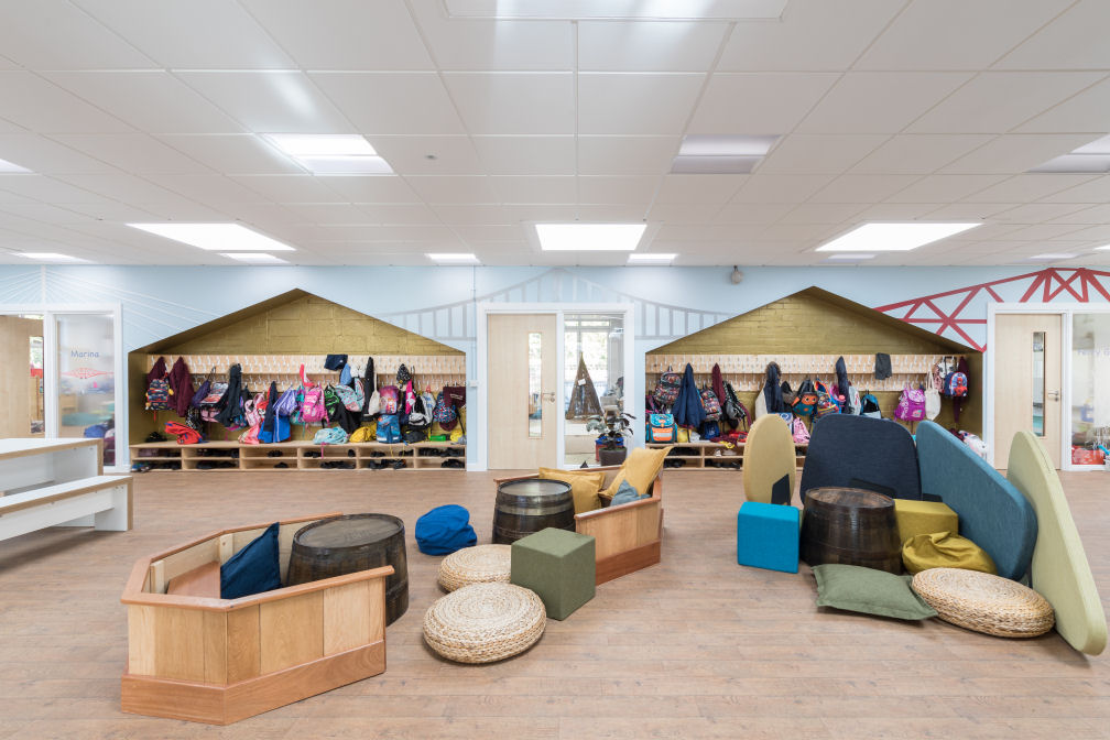 The cloakroom is shaped as a house, painted gold inside with coats and bags hanging on hooks and shoe storage beneath. on the walls is a graphic of the three Forth bridges. In the centre of the room there is a selection of yellow and blue upholstered seat cubes, beanbags and wicker seat pads scattered around three upturned barrels. Two wooden boat shaped benches for sitting. There are three acoustic dividers shaped like stones. There is a large white table with a bench at the rear of the room.