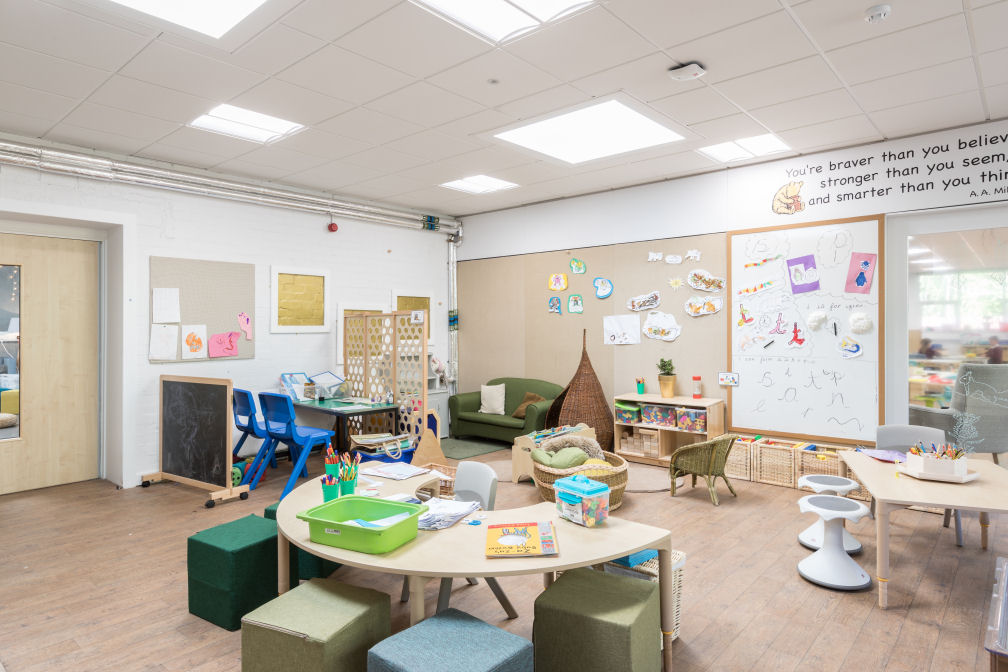 A white walled classroom with one wall covered in beige pin boarding fabric beside a white board. There is a U shaped table with a teacher chair on one side and four upholstered seating cubes in shades of green on the other side. There is a green sofa in the corner beside a small wicker nook. There are wooden storage units at low level along the back wall. A table with two wobble stools and a standard chair is to the right.