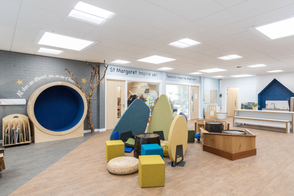 The sink area is recessed into a shape like a house, painted dark blue inside. On the wall over a circular nook it says "All stars are different, you are one of them!" there is a tree covered in fairy lights next to the nook. In the centre of the room there is a selection of yellow and blue upholstered seat cubes and wicker seat pads scattered around an upturned barrel. There are three acoustic dividers shaped like stones. On the wall above the classrooms are two saying "St Margaret inspire us!" and "teamwork makes the dream work". There is a large white table with a bench at the rear of the room.