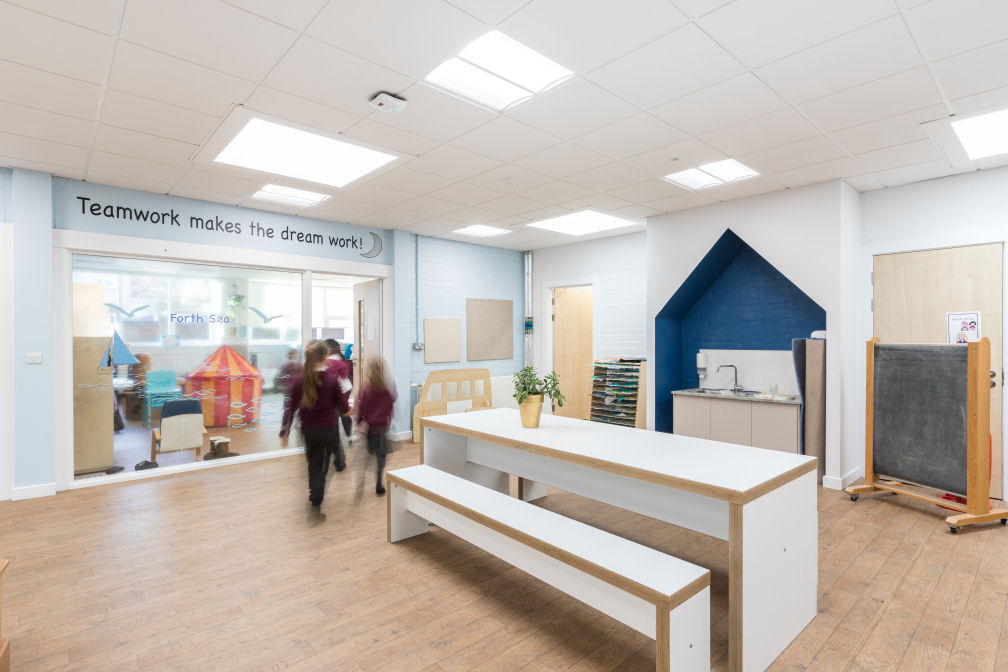 The sink area is recessed into a shape like a house, painted dark blue inside. On the wall above the classroom is a saying "teamwork makes the dream work". There is a large white table with a bench in the centre of the room. The door to the classroom has a big window to see in and out of the class, the window has graphics of a boat, fish and seagulls