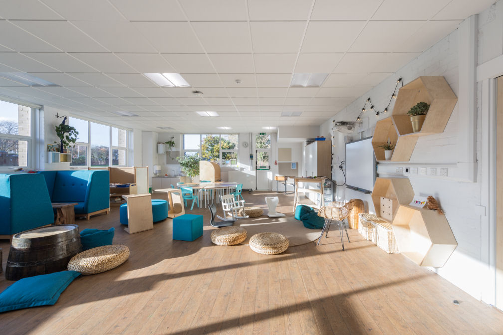 The Forth Sea classroom with lots of natural daylight, wood style flooring and white walls. The furniture is a mixture of natural wood tones and blue fabric. There are cushions, seat pads and upholstered cubes on the floor to sit on, a comfortable booth against the wall and a table with chairs in the background.