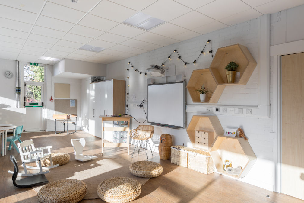 The Forth Sea classroom with lots of natural daylight, wood style flooring and white walls. The furniture is a mixture of natural wood tones. a wicker teacher chair in front of a Smart board, a couple of wicker baskets against the wall and an owl shaped wicker storage basket. In front of the chair is a circular rug surrounded by four wicker seat pads, two floor rocking seats and a traditional rocking chair. there are four hexagon shaped wooden shelves, two have plants inside, one has a small chest of drawers and the other has two cuddly toys. There are fairy lights hanging from the wall above the Smart board. A standing teachers desk is next to the Smart board in front of two cupboards.