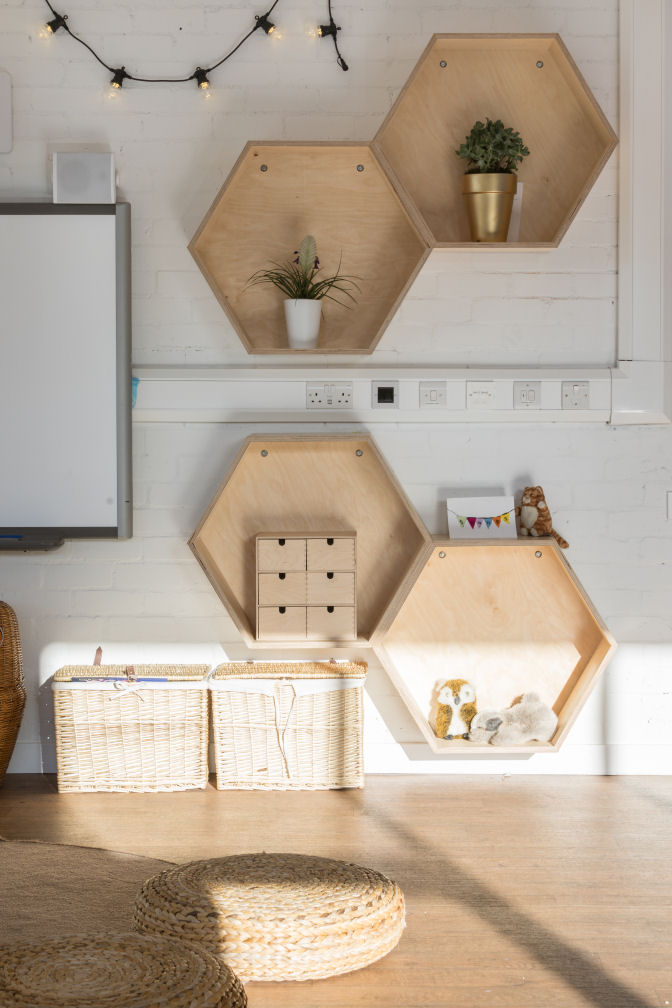 A close up of the storage units on the wall. there are four hexagon shaped wooden shelves, two have plants inside, one has a small chest of drawers and the other has two cuddly toys.