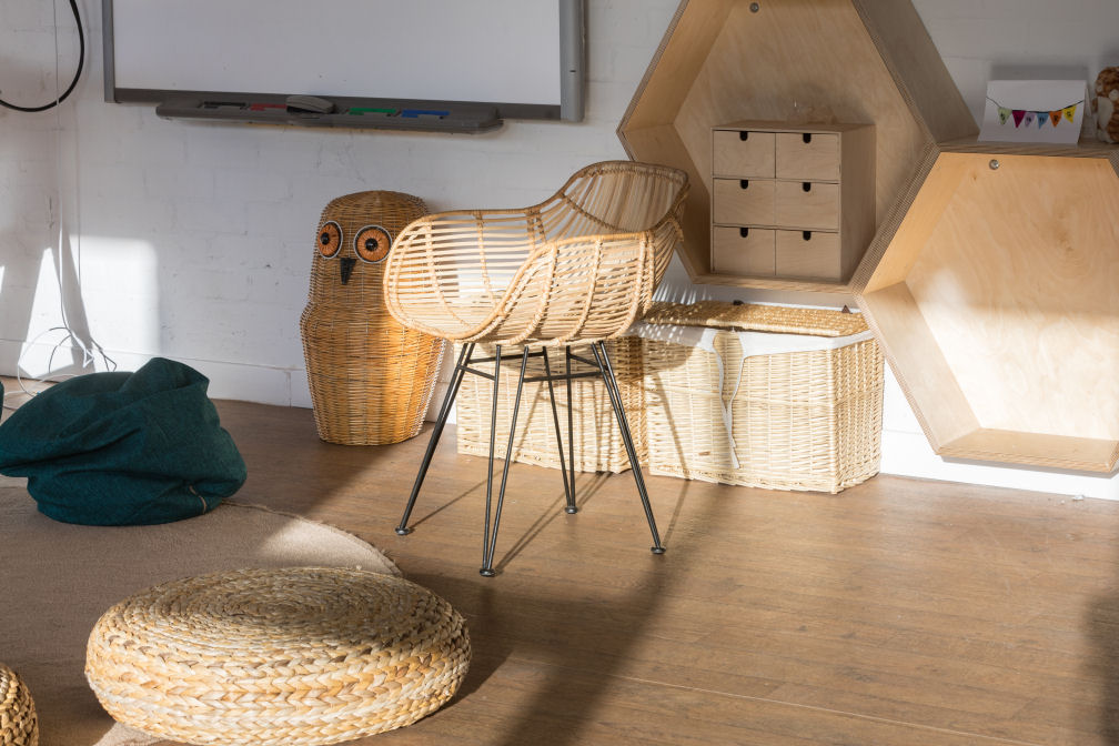 A close up of a wicker teacher chair in front of a Smart board, a couple of wicker baskets against the wall and an owl shaped wicker storage basket. In front of the chair is a circular rug with a blue bean bag and wicker seat pad.