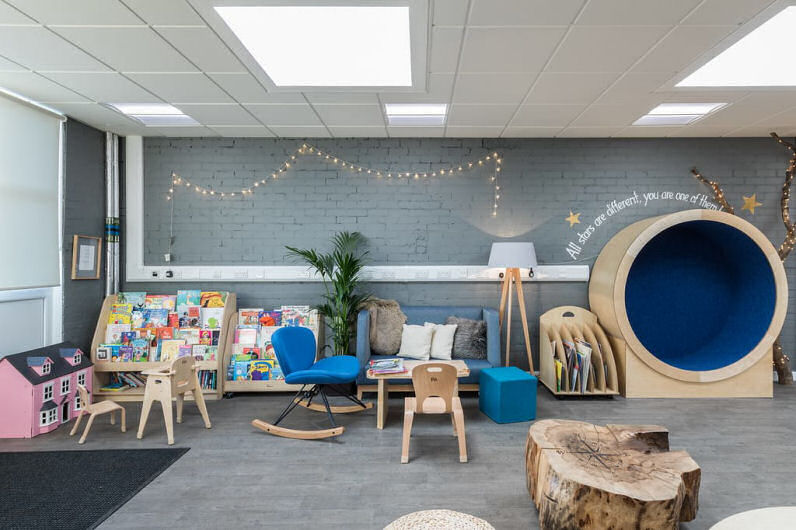 the general purpose room has a library corner which is like a living room, a circular nook is against the wall for a child to sit in, a sofa and rocking chair are beside a coffee table and a lamp and plant create a homely look