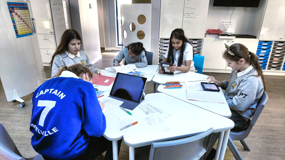 five girls sit at a hexagonal table. two are working on ipads and three on paper
