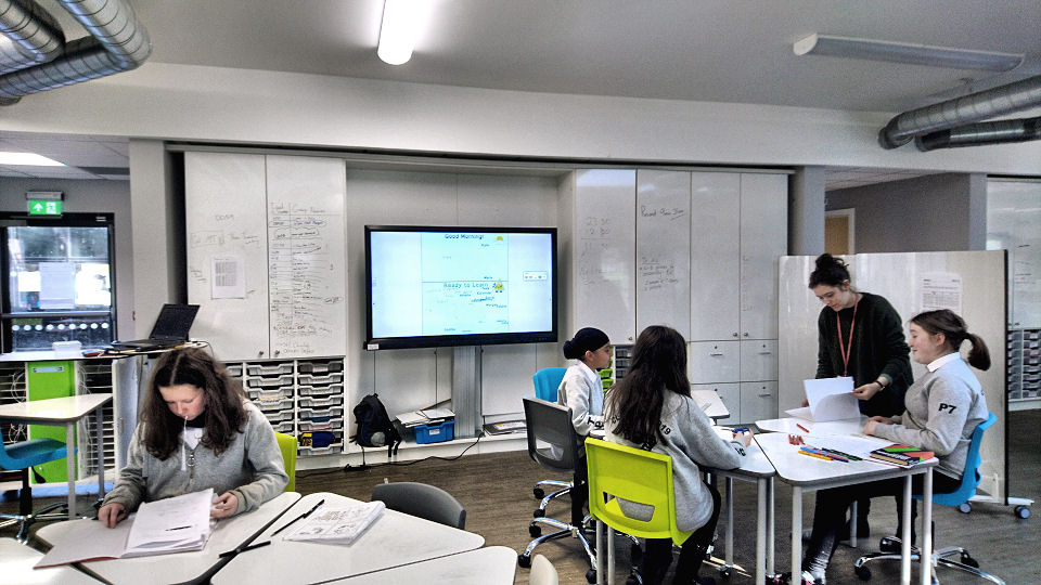 three children talk to their teacher, the TV screen has work on it and the whiteboard cupboards have notes on them