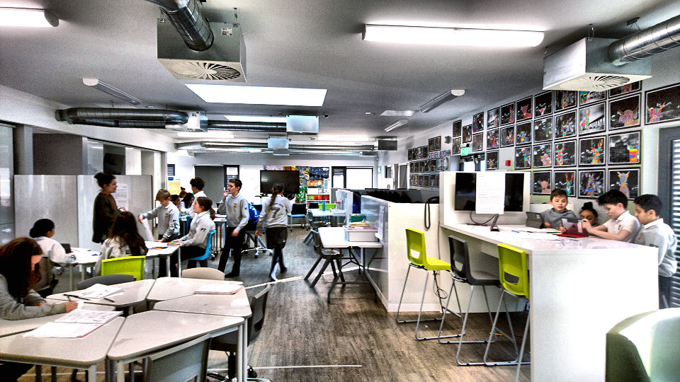 the length of the classroom, on the left are groupings of triangular tables and on the right is a high standing workbench with high stools