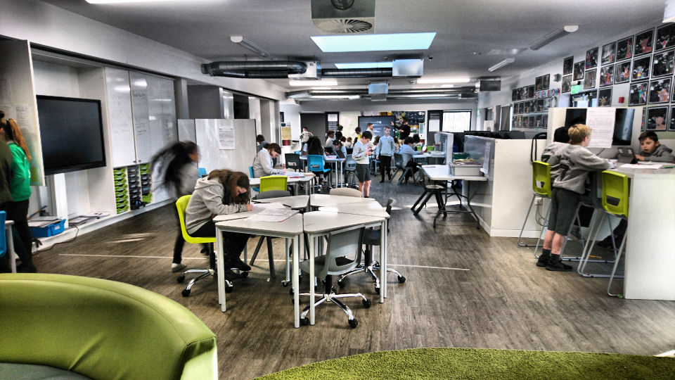 the children work in different working zones in the room. a boy stands at the standing table beside two boys who are sitting on the high chairs. other children are sitting at triangular tables