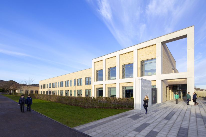 The front of the school, there is a pavement and grass up to a wall, children are walking to the building
