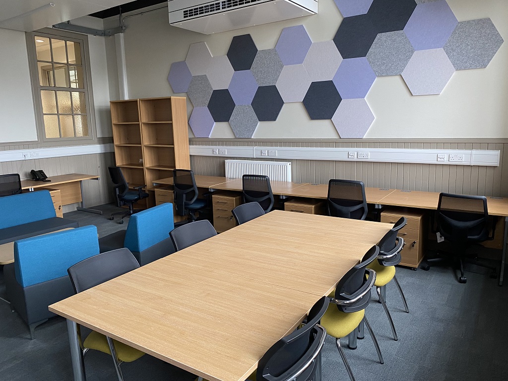 A classroom with acoustic hexagons on the wall providing a colourful design feature, a rectangular table with chairs, some wide upholstered seats around a coffee table and benching against the wall with wheelie chairs