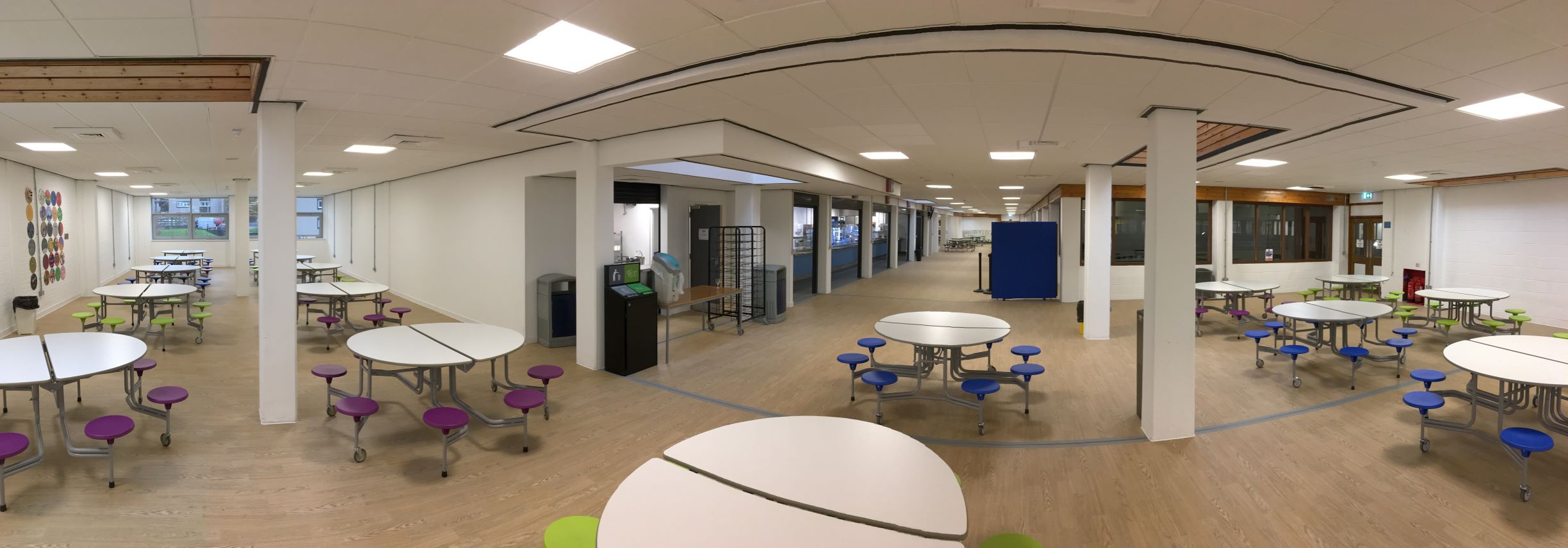 A large room with multiple circular dining tables with attached colourful round stools on wheels